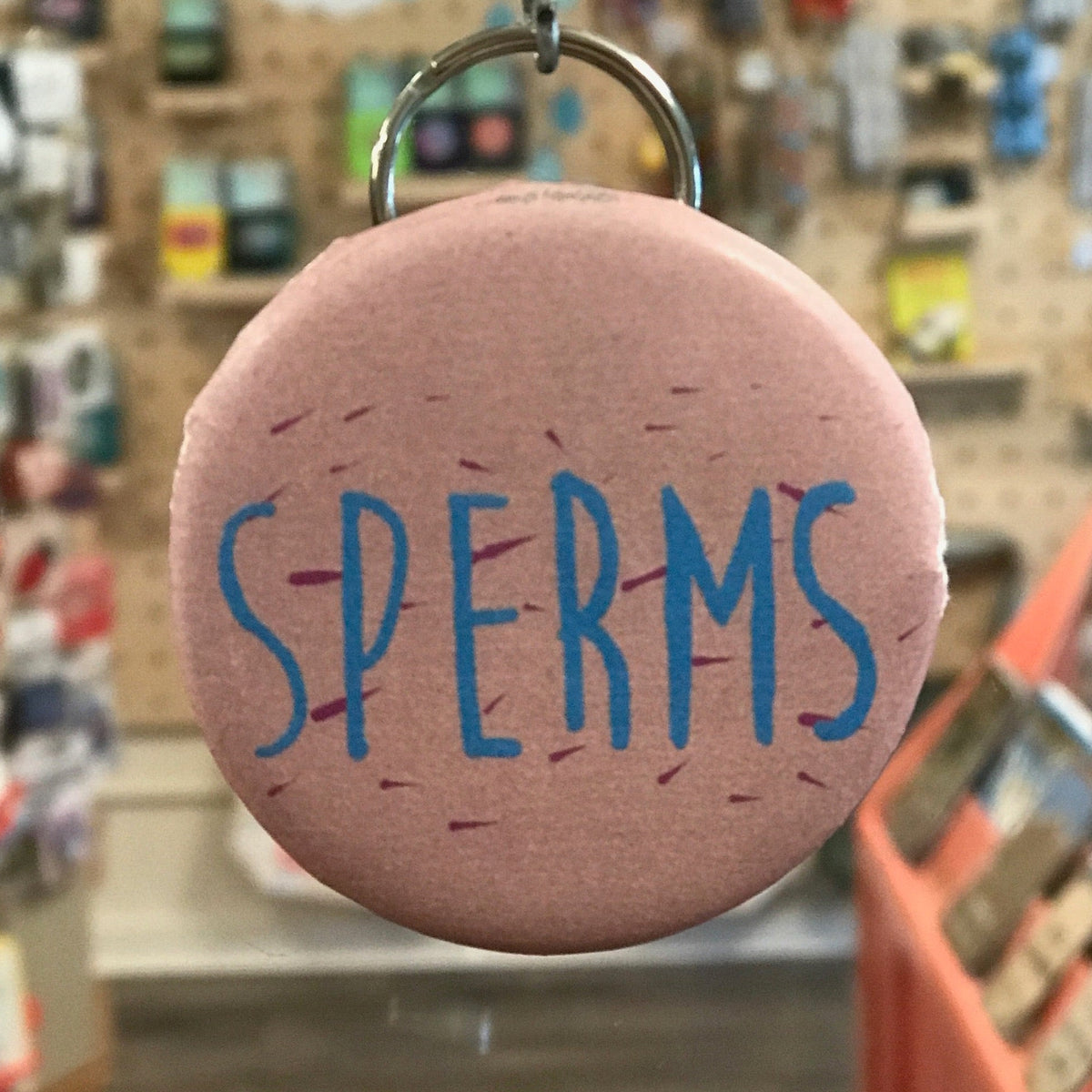 Pink keychain with 'SPERMS' text in blue on a store shelf background
