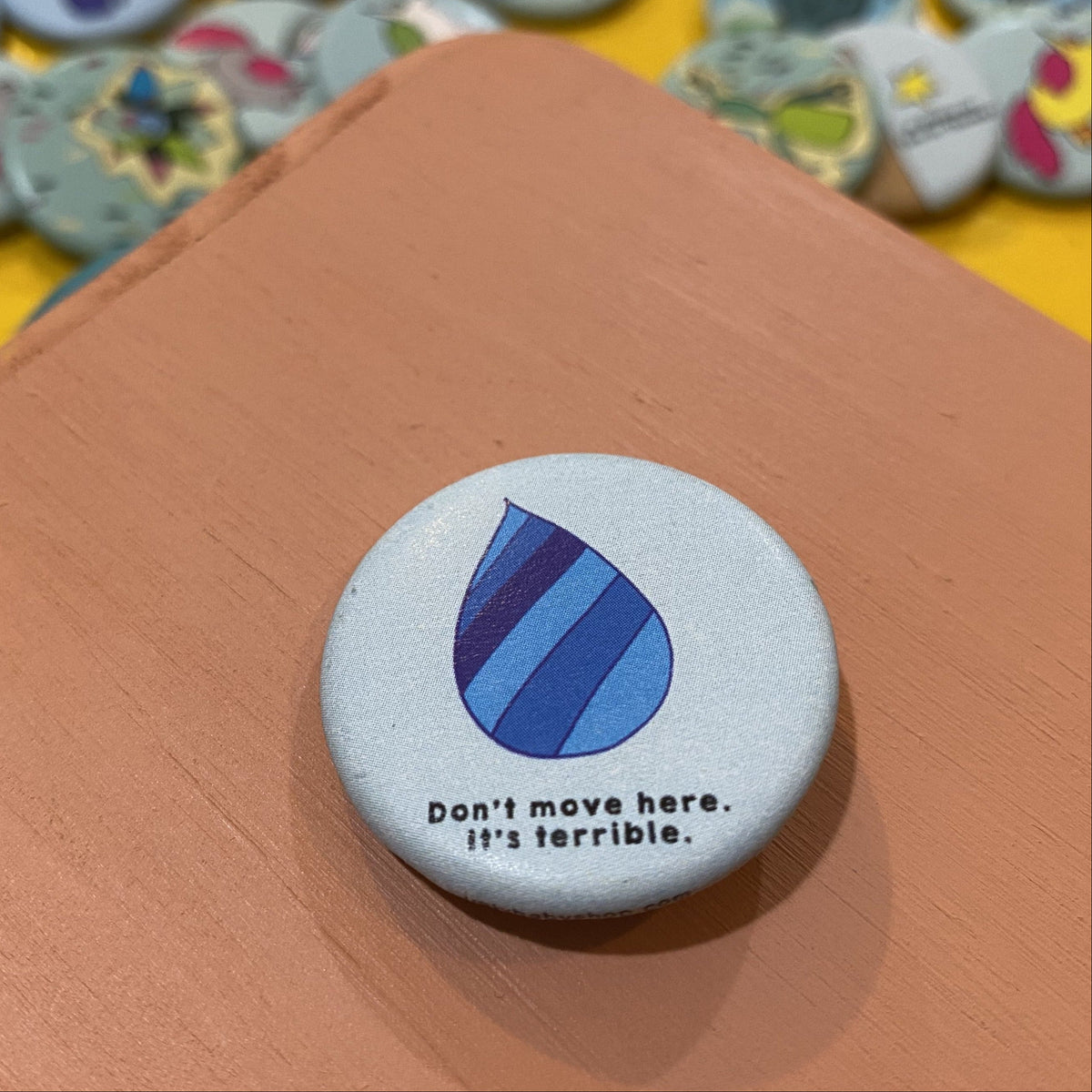 Button badge with blue droplet design and text 'Don't move here, it's terrible' on a wooden surface with other similar badges in the background.