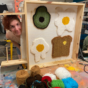 Person standing next to a wooden tufting frame with yarn art of an avocado, eggs, and toast.