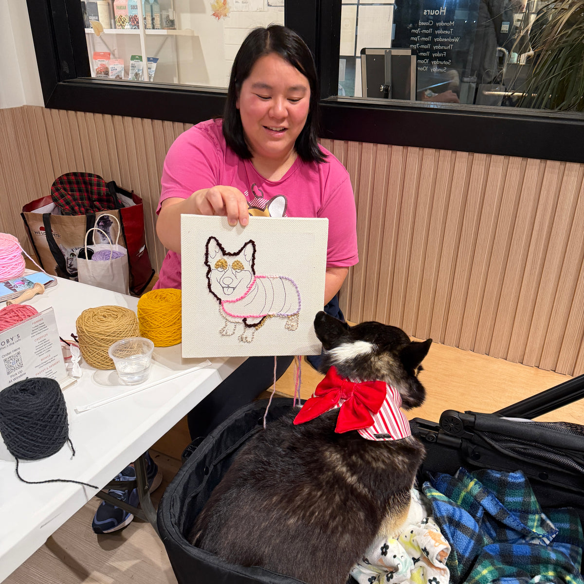 Woman holding a punch needle art piece of a dog, sitting next to a dog wearing a red bow in a casual setting.