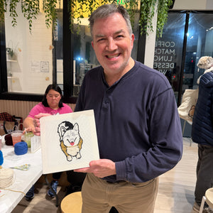 Man holding a punch needle portrait of a dog at an event with other people and tables in the background.