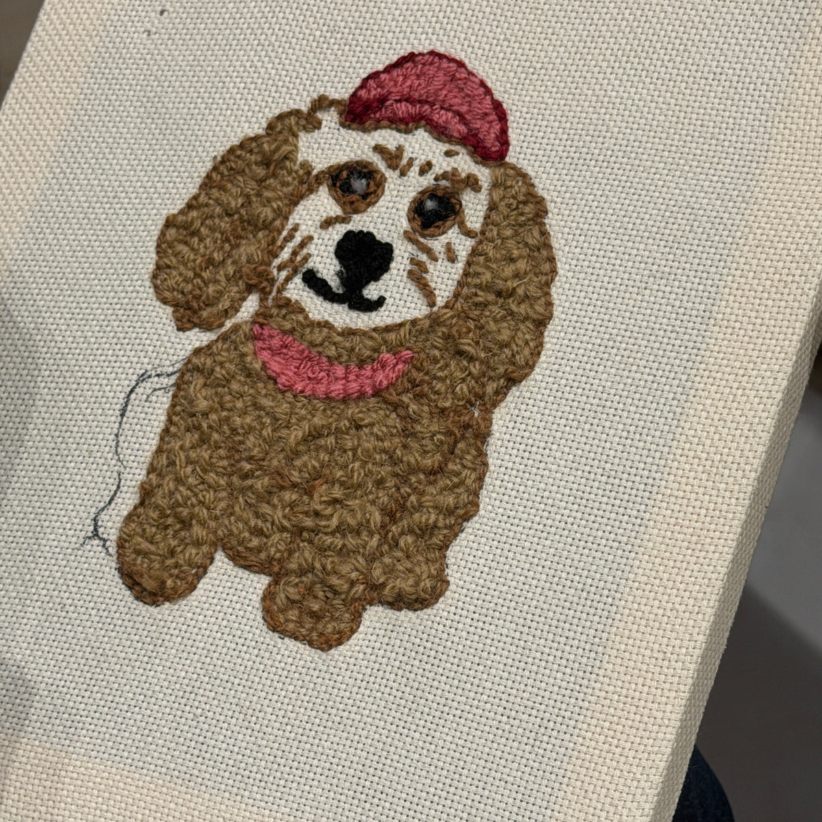 Partially finished punch needle pet portrait with a pink hat on beige fabric. 