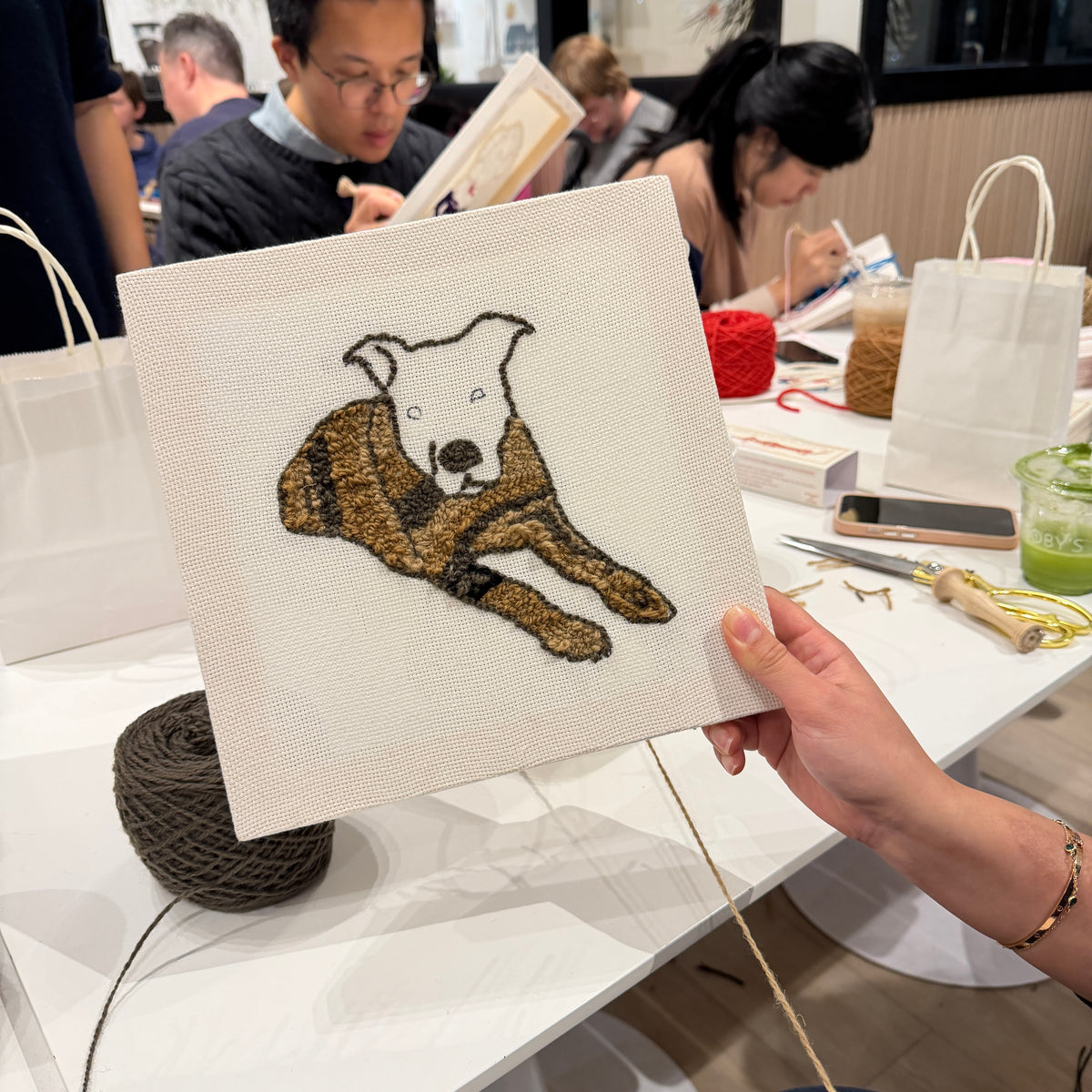Person holding a punch needle pet portrait of a dog at an event with other attendees.