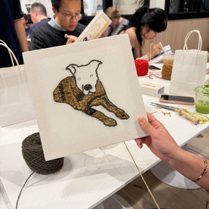 Person holding a punch needle pet portrait of a dog at an event with other attendees.