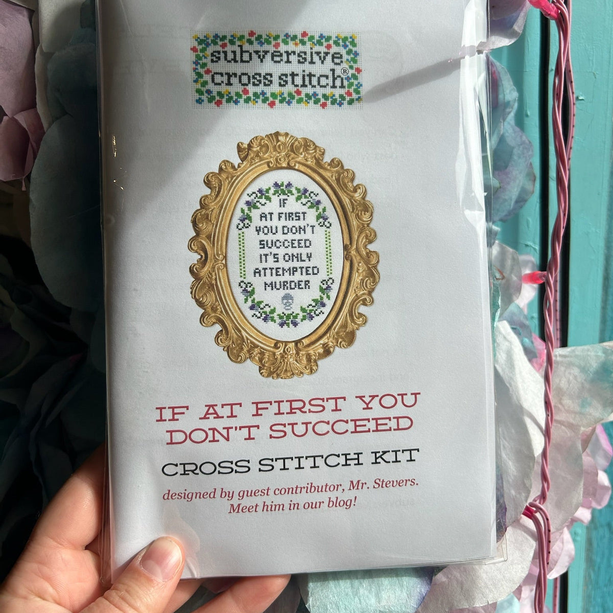 Cross stitch kit packaging held by a hand with decorative background. The kit says, "If at first you don't succeed it's only attempted murder." 