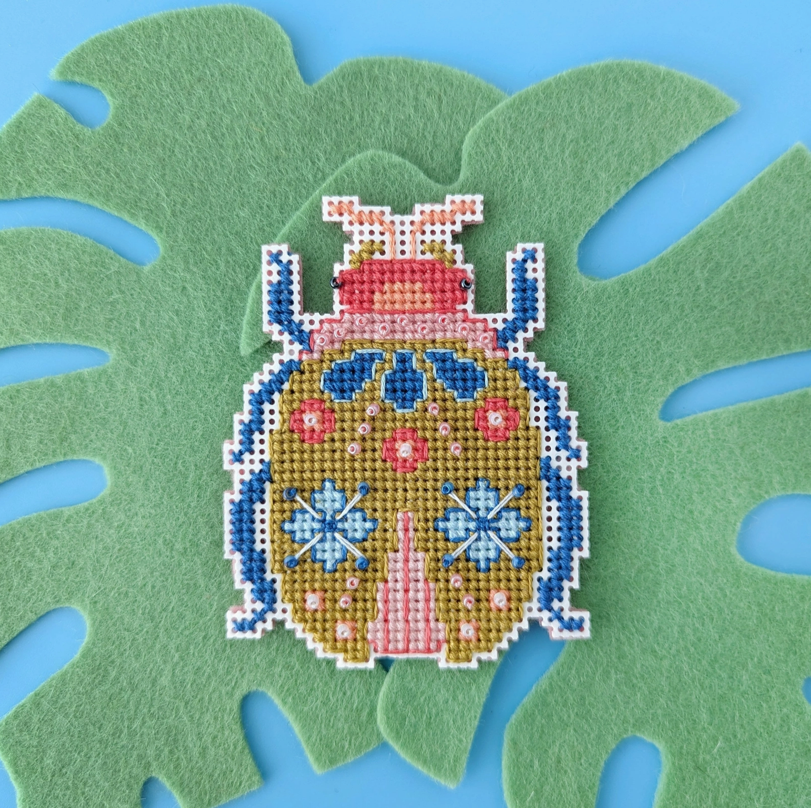 A cross stitch ornament of a beetle is sitting atop a felt Monstera leaf. 