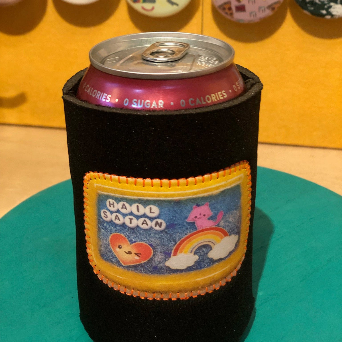 A black foam can cooler sleeve holding a pink beverage can, with a colorful front patch reading 'HAIL SATAN' and showing cartoon hearts, a rainbow, and a pink character. The sleeve sits on a teal round board against a yellow wall with decorative pins behind it.
