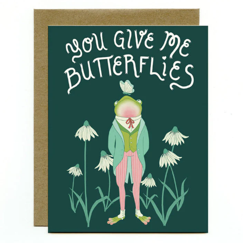 A green card with the words "You give me butterflies" and an illustration of a frog wearing a suit with a butterfly on his nose standing in a field of daisies.