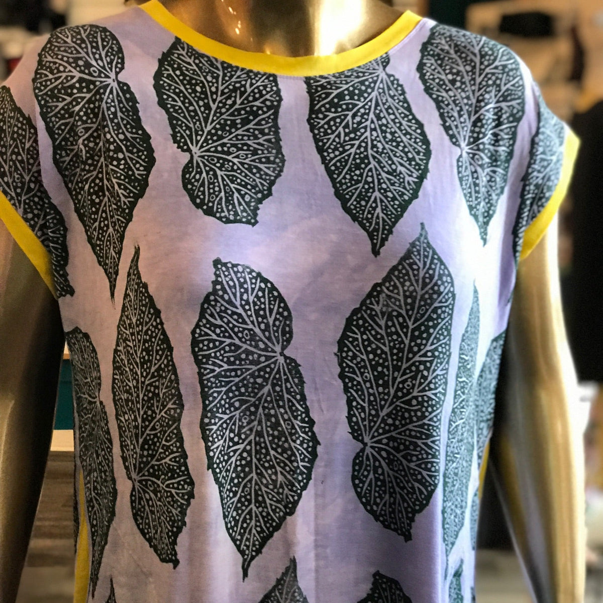 Mannequin wearing a sleeveless top with begonia leaf pattern printed in forest green on a light purple background. 