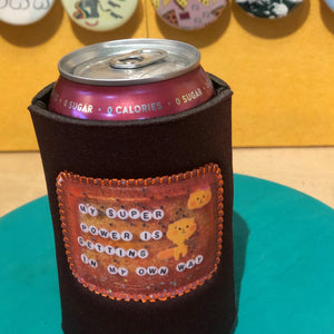 A black neoprene can cooler sleeve holding a pink can, displayed on a teal circular surface with a yellow wall and decorative pins in the background.