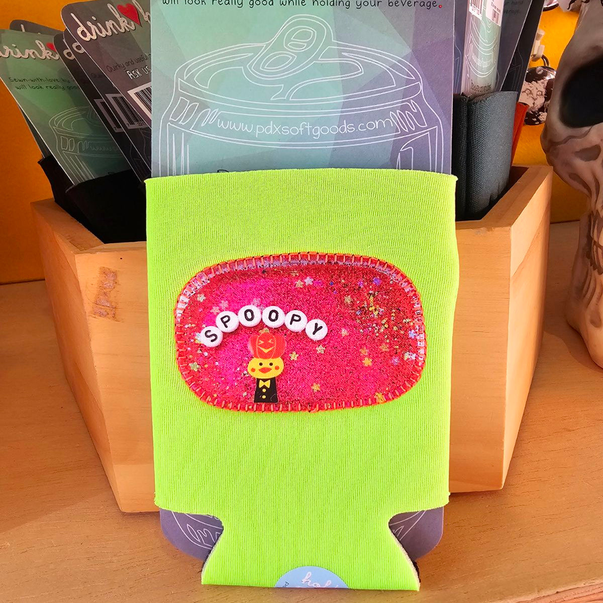 A lime green drink holder with a pink glittery 'Spoopy' patch featuring a small cartoon character, displayed in a wooden holder with other packaged items behind it.