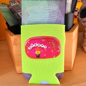 A lime green drink holder with a pink glittery 'Spoopy' patch featuring a small cartoon character, displayed in a wooden holder with other packaged items behind it.