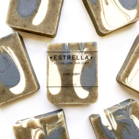 A collection of Estrella all natural bar soaps, with a bar labeled 'Earl Grey' with a swirl of gray and white color on it, indicating the soap is designed with a visual appeal of Earl Grey tea leaves and speckles.