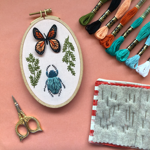 Embroidery hoop with butterfly and beetle design, embroidery threads, scissors, and fabric on a pink background. 