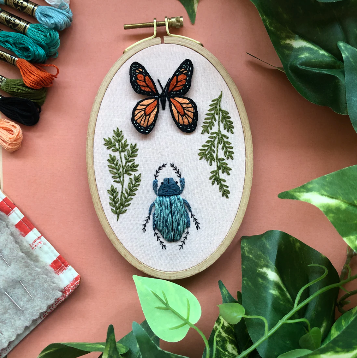 Embroidery hoop with butterfly and beetle design on a pink background with embroidery threads and plants.