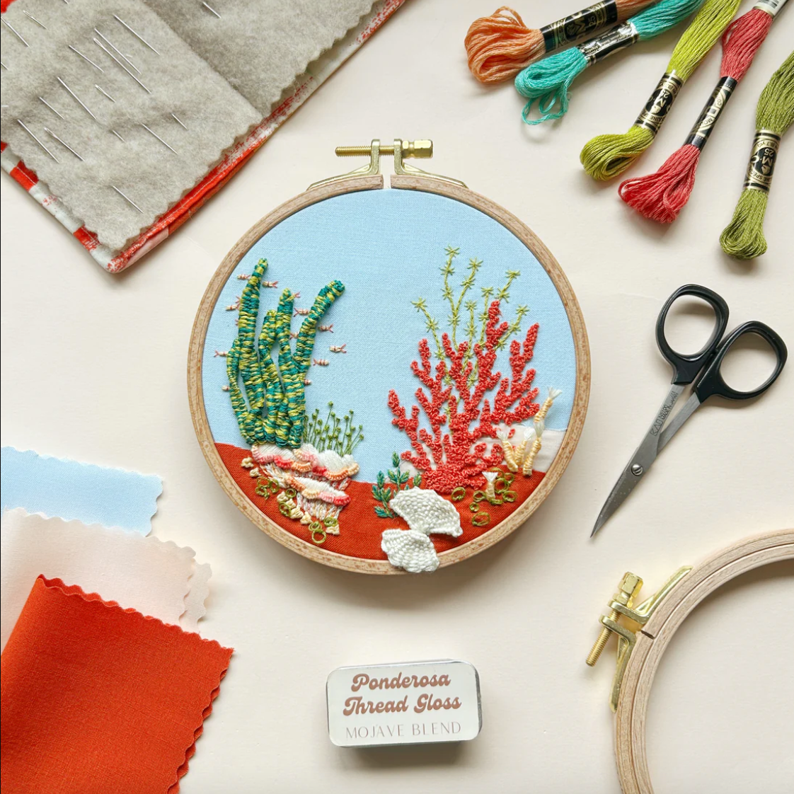 Embroidery hoop with underwater scene, thread spools, scissors, and fabric on a light background. 
