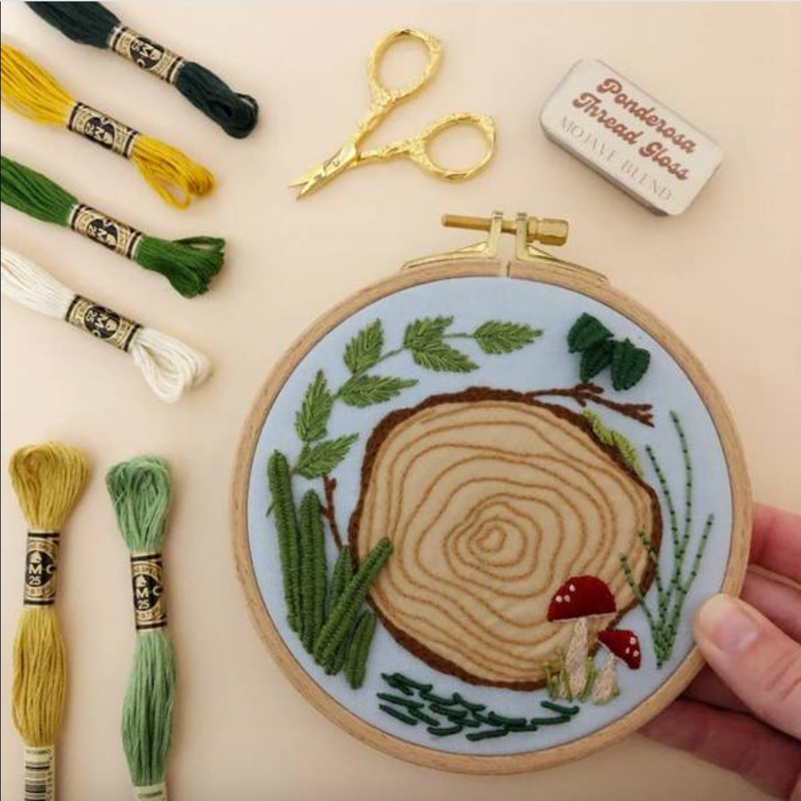 Embroidery hoop with tree ring design surrounded by embroidery threads, scissors, and a thread gloss on a beige background.
