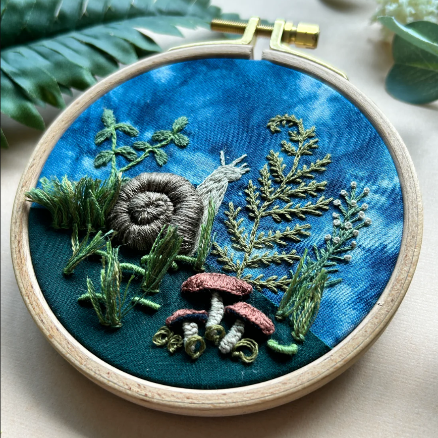 Embroidery hoop with embroidered scene of plants and a snail on blue fabric.
