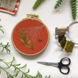 Embroidery hoop with spider web and leaf design on red fabric, surrounded by greenery and embroidery tools.