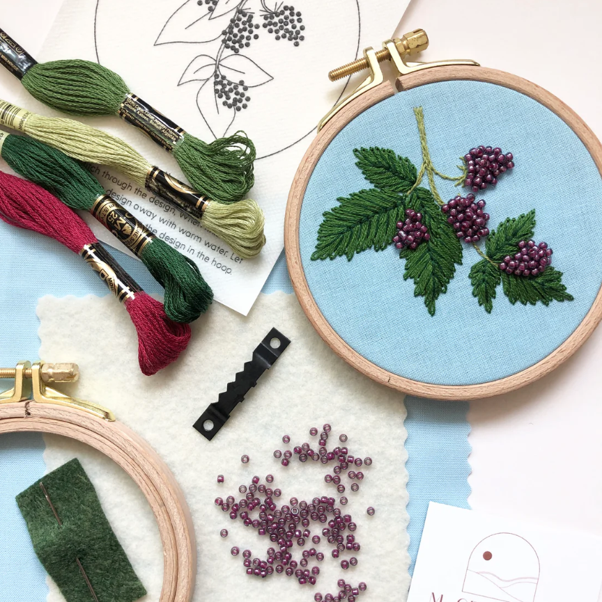 Embroidery hoop with blackberry design and embroidery threads on a light blue background.