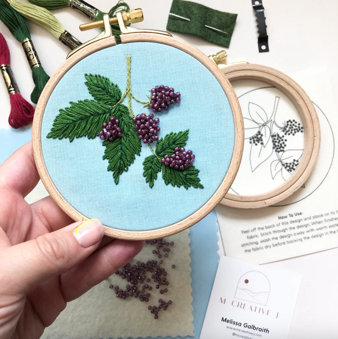Embroidery hoop with blackberry design held by a hand, surrounded by embroidery materials on a light blue surface.