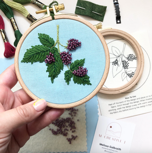 Embroidery hoop with blackberry design held by a hand, surrounded by embroidery materials on a light blue surface.