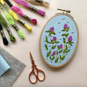 Embroidery hoop with clover design, embroidery threads, scissors, and fabric on a light background. 