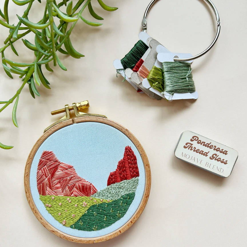 Embroidery hoop with mountain design, keychain, and thread package on a light background. 
