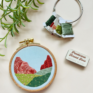 Embroidery hoop with mountain design, keychain, and thread package on a light background. 