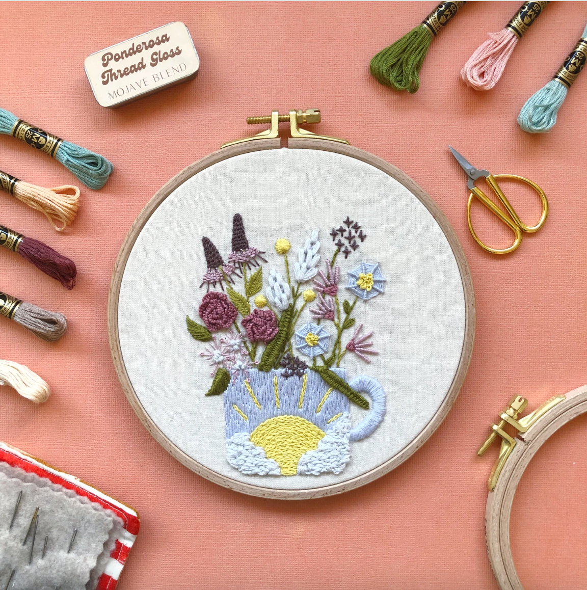Embroidery hoop with floral design on a pink background with embroidery threads and scissors.