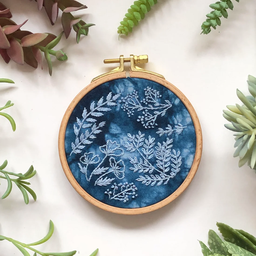 Embroidery hoop with blue fabric and white floral pattern, surrounded by leaves on a light background. 