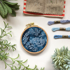 Embroidery hoop with floral design on a white surface with embroidery threads and plants.
