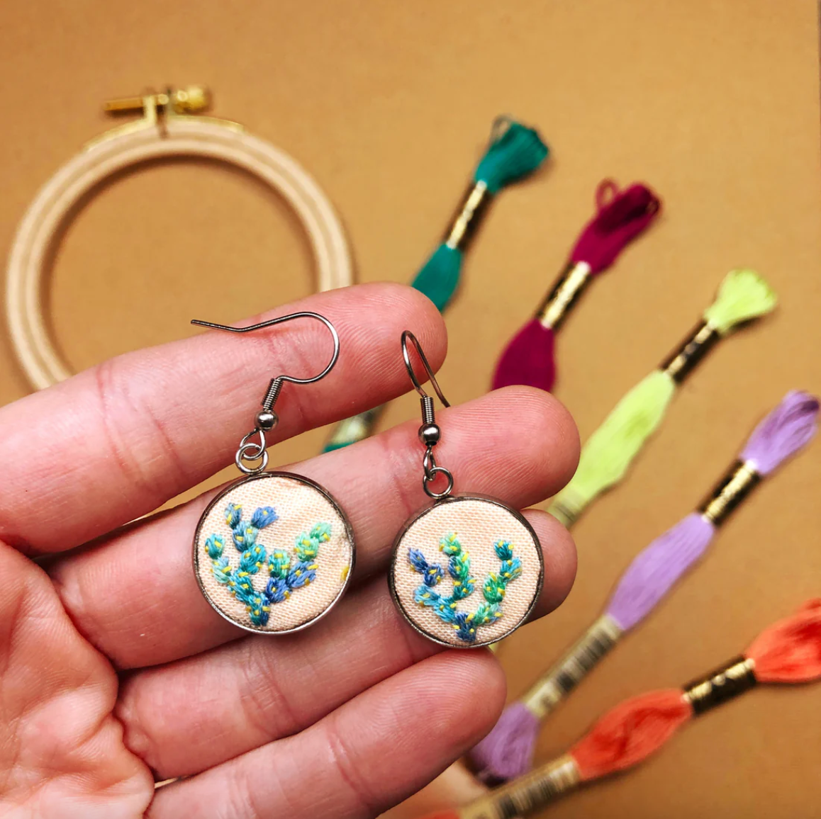 Hand holding embroidered earrings with colorful thread spools in the background. 