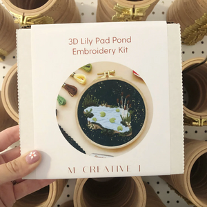 3D Lily Pad Pond Embroidery Kit packaging held by a hand with a blurred background of embroidery hoops.