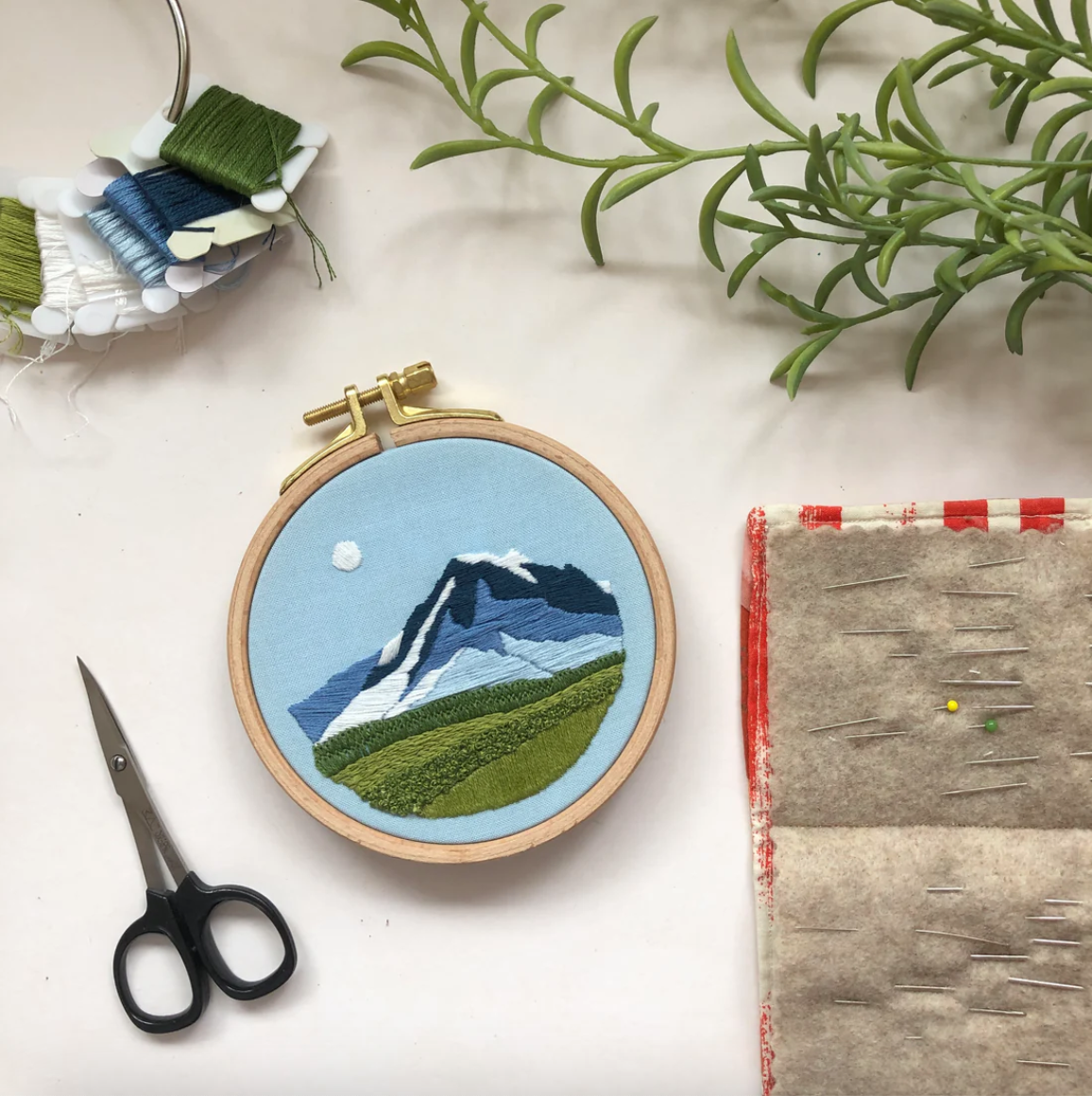 Embroidery hoop with mountain design on a white surface with sewing tools and plants.