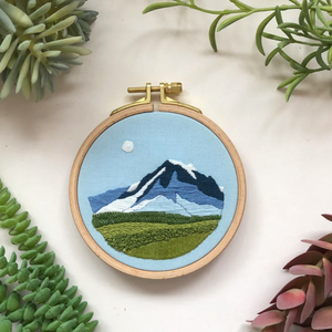 Embroidery hoop with mountain design surrounded by green plants on a white background. 