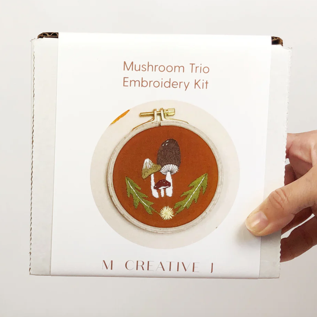 Mushroom Trio Embroidery Kit held by a hand on a light gray background. 