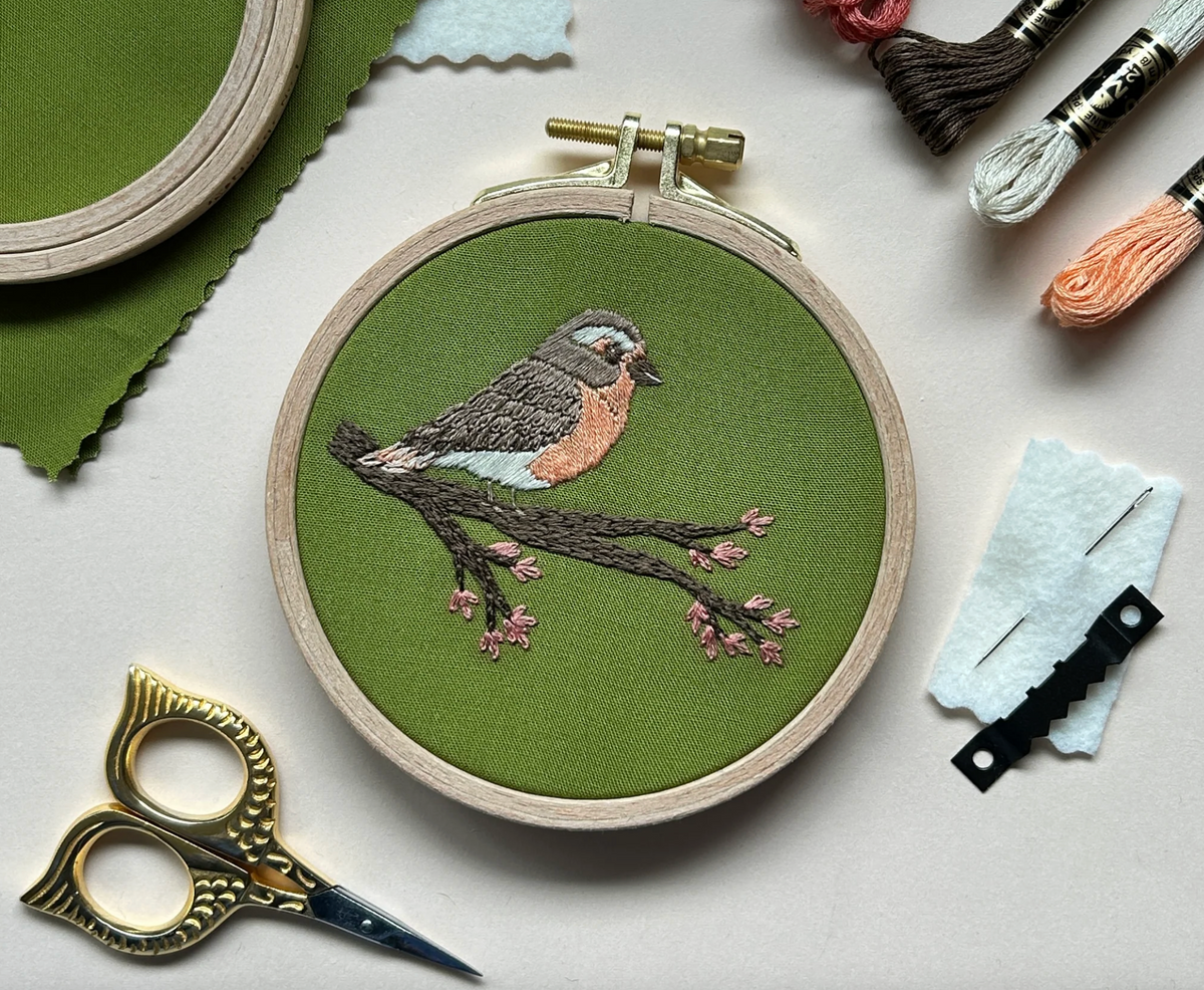 An embroidered robin sits on a flowering branch against a green fabric background. The embroidered piece is surrounded by embroidery floss, needles, a sawtooth hanger and little gold owl embroidery scissors. 