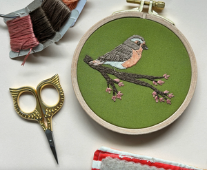 An embroidered robin sits on a flowering branch against a green fabric background. The embroidered piece is surrounded by embroidery floss, needles, a sawtooth hanger and little gold owl embroidery scissors. 