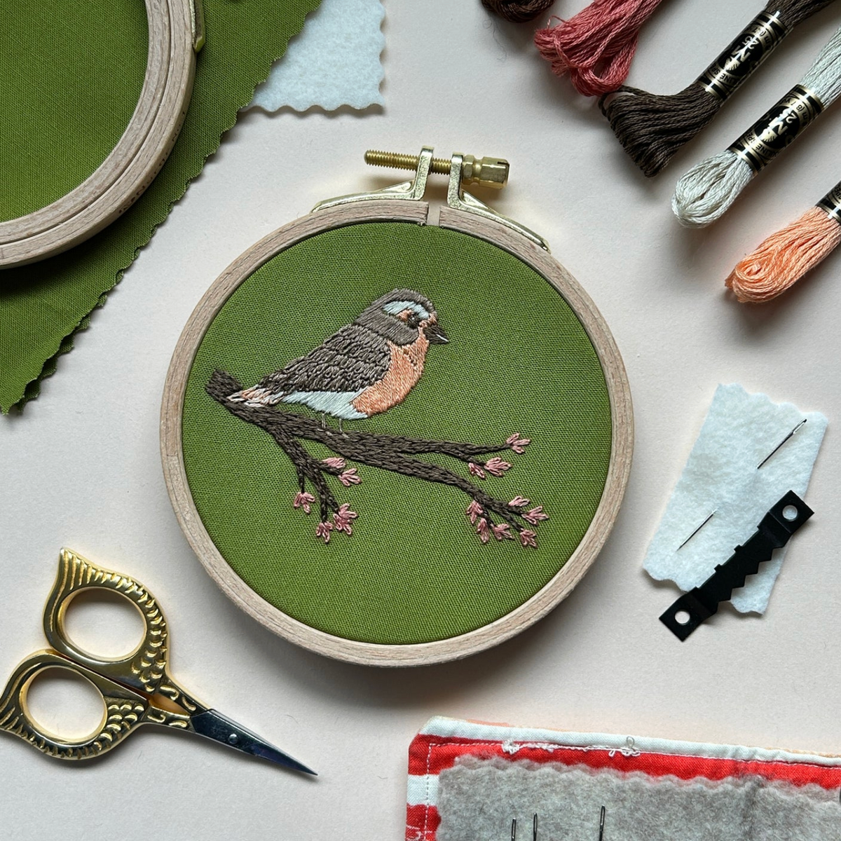 An embroidered robin sits on a flowering branch against a green fabric background. The embroidered piece is surrounded by embroidery floss, needles, a sawtooth hanger and little gold owl embroidery scissors. 