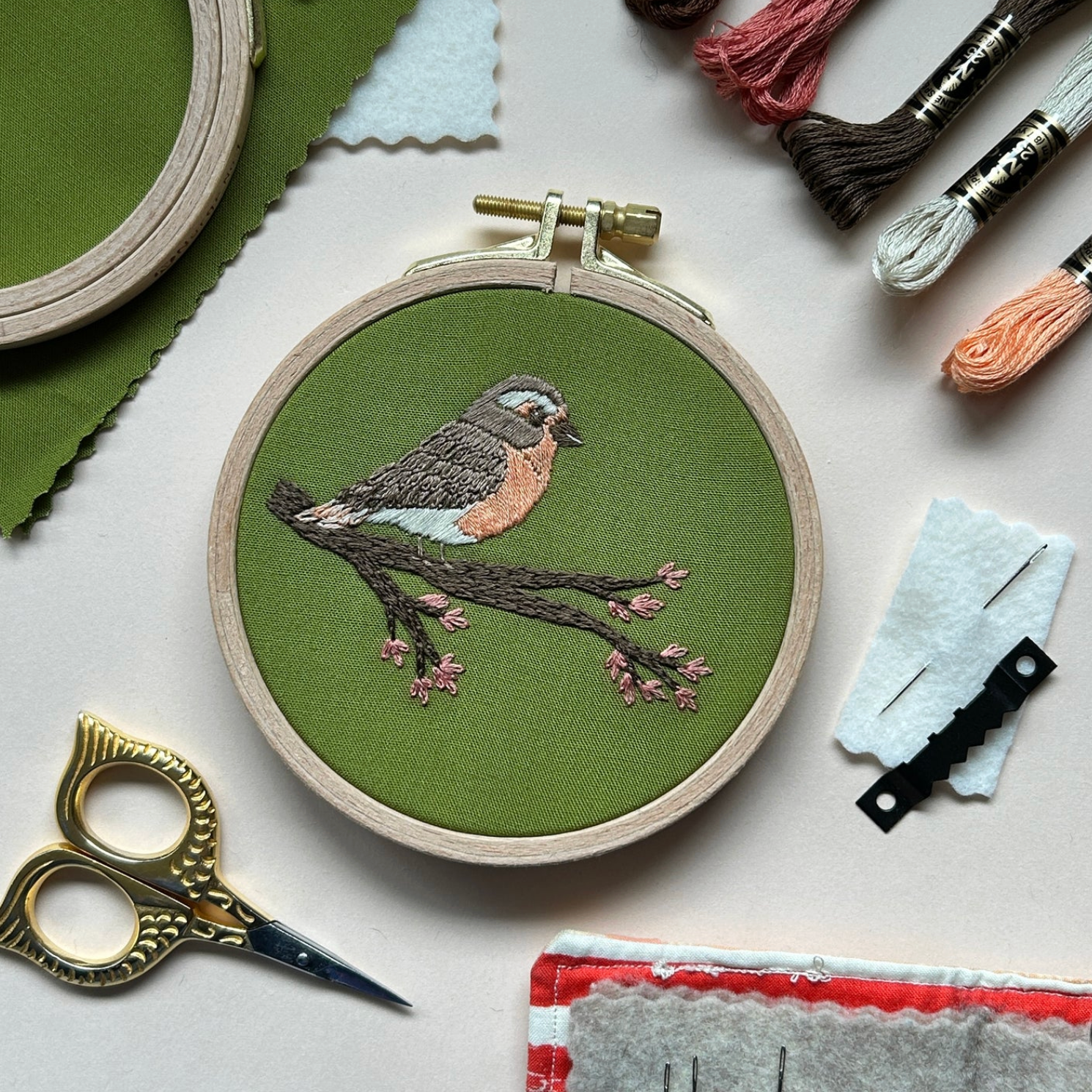 An embroidered robin sits on a flowering branch against a green fabric background. The embroidered piece is surrounded by embroidery floss, needles, a sawtooth hanger and little gold owl embroidery scissors. 