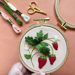 Embroidery hoop with strawberry design on a pink background with embroidery tools.