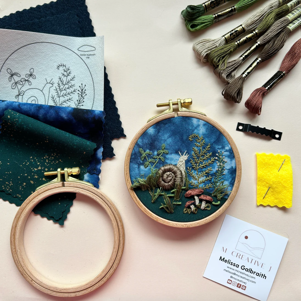 Embroidery hoop with snail design, fabric swatches, and embroidery tools on a light surface.