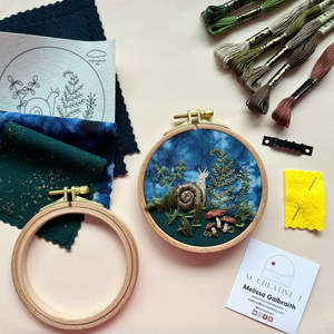 Embroidery hoop with snail design, fabric swatches, and embroidery tools on a light surface.