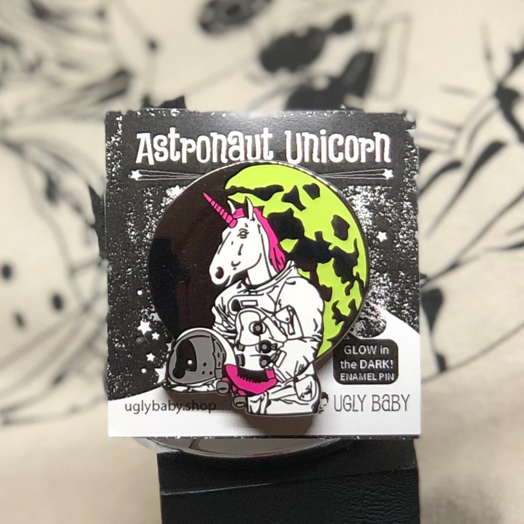 An enamel pin of an astronaut unicorn on its original backing card, photographed against a black and white patterned background.