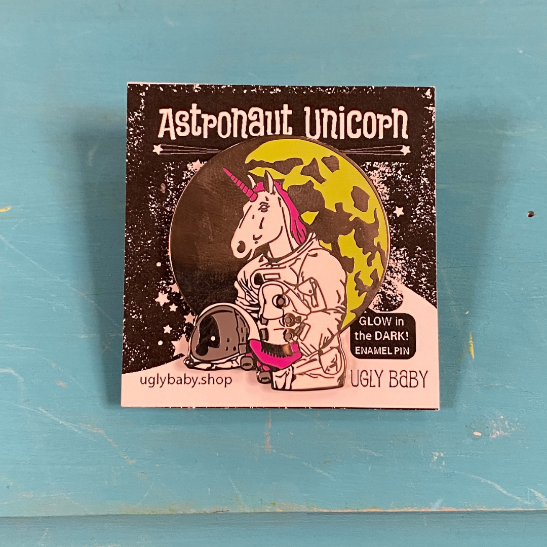 An enamel pin of an astronaut unicorn with a pink horn and tail, set against a moon and displayed on its original backing card. The card features the text 'Astronaut Unicorn' and the brand 'Ugly Baby'.
