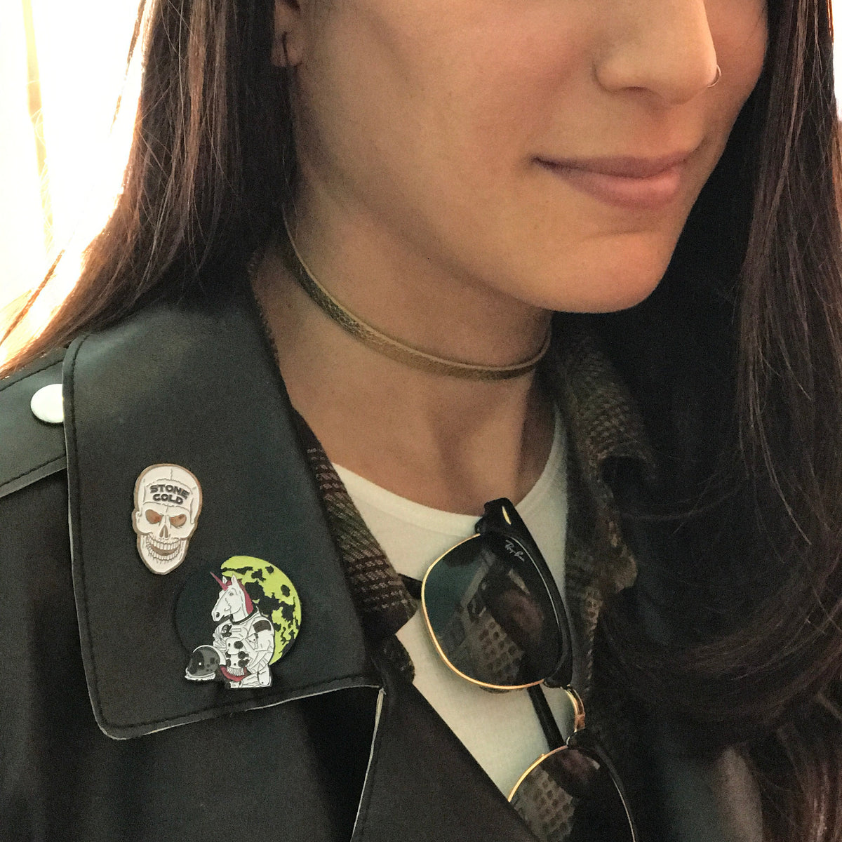 A close-up portrait of a woman wearing a black leather jacket adorned with two unique enamel pins on the collar, including a skull pin and an astronaut unicorn with a green moon background.