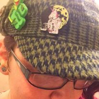 A close-up of a person wearing a green and black houndstooth flat cap with decorative pins, glasses, and a pink top.