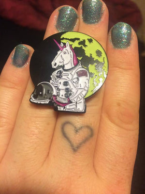 A close-up of a hand with glittery green nail polish holding an enamel pin. The pin features a white astronaut unicorn with a pink horn, holding a small skull, set against a black background and a yellow-green planet.