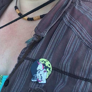 A close-up selfie of a person outdoors wearing a brown and gray vertically striped button-up shirt, a black cord necklace, and a colorful Astronaut Unicorn on the chest.